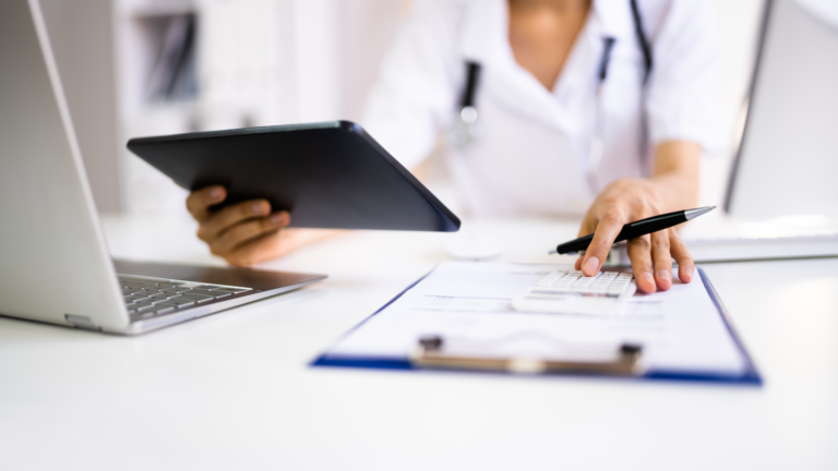 A doctor looking at a clipboard and tablet in front of a ocmputer